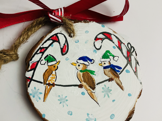 painted wood ornament shows three little birds sitting on a wire. The wire is attached to two candy canes that are red and white. Little brown birds wear holiday blue and green hats and scarfs. Snowflakes are white and light blue in the background.